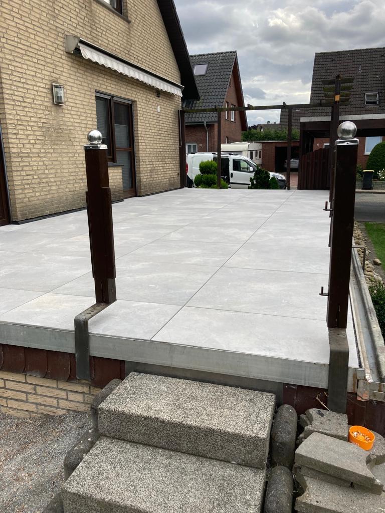 Wykończony taras z szarych płyt betonowych, schody z szarego kamienia, widok na dom z cegły i biały samochód dostawczy w tle.