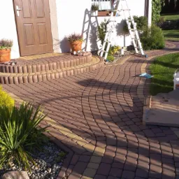 Kaskadowe schody wejściowe i ścieżka z kostki brukowej w odcieniach brązu i żółci, otoczone zielenią, z dwoma wiadrami i kartonem na trawniku, drabina oparta o ścianę domu w tle.