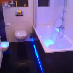 Nowoczesna łazienka z czarną podłogą, białymi płytkami i niebieskim oświetleniem LED pod wanną i w umywalce.