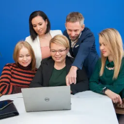 Zesp&oacute;ł pięciu os&oacute;b pochylony nad laptopem Dell na białym stole, w tle niebieska ściana i roślina doniczkowa. Mężczyzna w granatowej marynarce wskazuje palcem na ekran. Na stole leżą telefon...