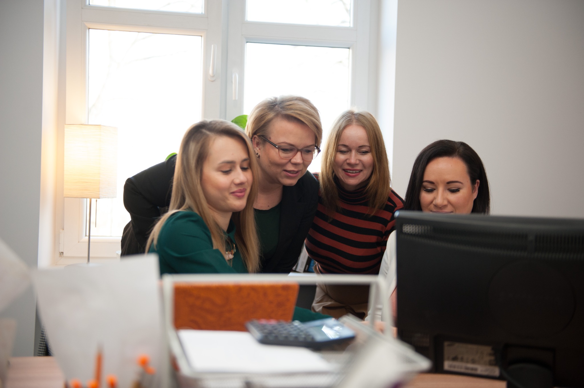 Cztery kobiety w biurze, pochylone nad monitorem komputera, analizują dane, na biurku kalkulator i dokumenty.