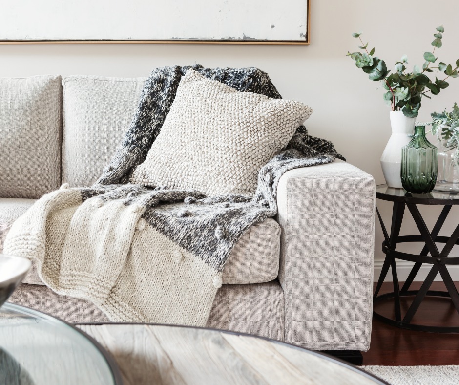 Jasnoszara sofa z dekoracyjną poduszką o grubej fakturze i dzierganym pledem w odcieniach szarości; obok stolik z metalową podstawą i wazonami z zielenią, całość w jasnym, minimalistycznym wnętrzu.