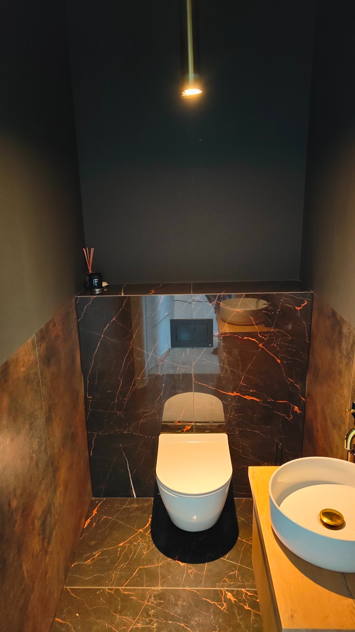 Nowoczesna łazienka z ciemnymi ścianami i podłogą z marmurowymi żyłami, podwieszana toaleta, okrągła umywalka nablatowa i minimalistyczna lampa sufitowa.