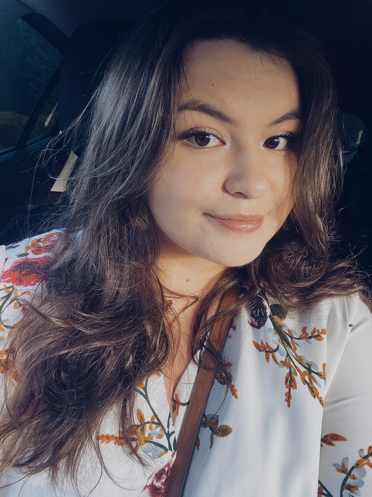 Portret kobiety z długimi brązowymi włosami, w bluzce w kwiaty, z delikatnym makijażem, siedzącej w samochodzie.