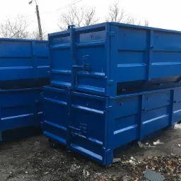 Stos niebieskich, metalowych kontenerów na podmokłym placu budowy, widoczne elementy mocujące i łańcuchy.