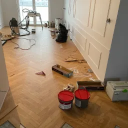 Układanie parkietu w jodełkę: widok na fragment podłogi w trakcie montażu, z widocznymi narzędziami, klejem SMP Elastic 300 w czerwonych wiaderkach, niezapakowanymi deskami, odkurzaczem...