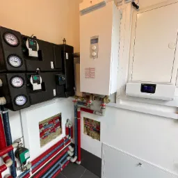 Węzeł cieplny z dwoma obiegami grzewczymi, widoczne pompy obiegowe, manometry, zawory oraz rury w izolacji czerwono-niebieskiej. Na ścianie biały piec grzewczy i regulator temperatury.