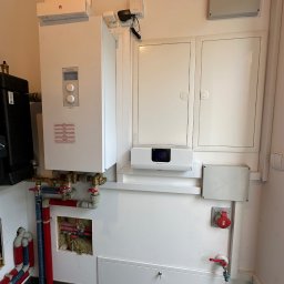 Usługi budowlane Majerczyk - Biała instalacja grzewcza Kospel z widocznymi rurami, zaworami, czarnym zbiornikiem, skrzynkami elektrycznymi i routerem Huawei na górze, zamontowana na białej ścianie w pomieszczeniu technicznym.