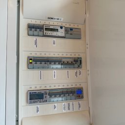 Usługi budowlane Majerczyk - Otwarta skrzynka rozdzielcza z bezpiecznikami i licznikami energii elektrycznej, etykiety opisujące obwody, widoczne marki Hager, Orno i Eastron.