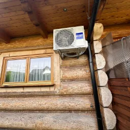 Biała jednostka zewnętrzna klimatyzacji Haier zamontowana na drewnianej ścianie domu z bali, widoczna rynna spustowa i okno z firankami.