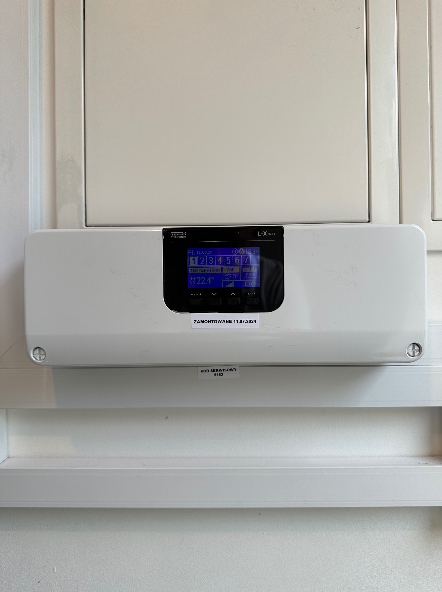 Biały sterownik klimatyzacji TECH L-X WiFi zamontowany na ścianie, z wyświetlaczem pokazującym temperaturę 22.4°C, datą 12.07.24 i kodem serwisowym 5162.