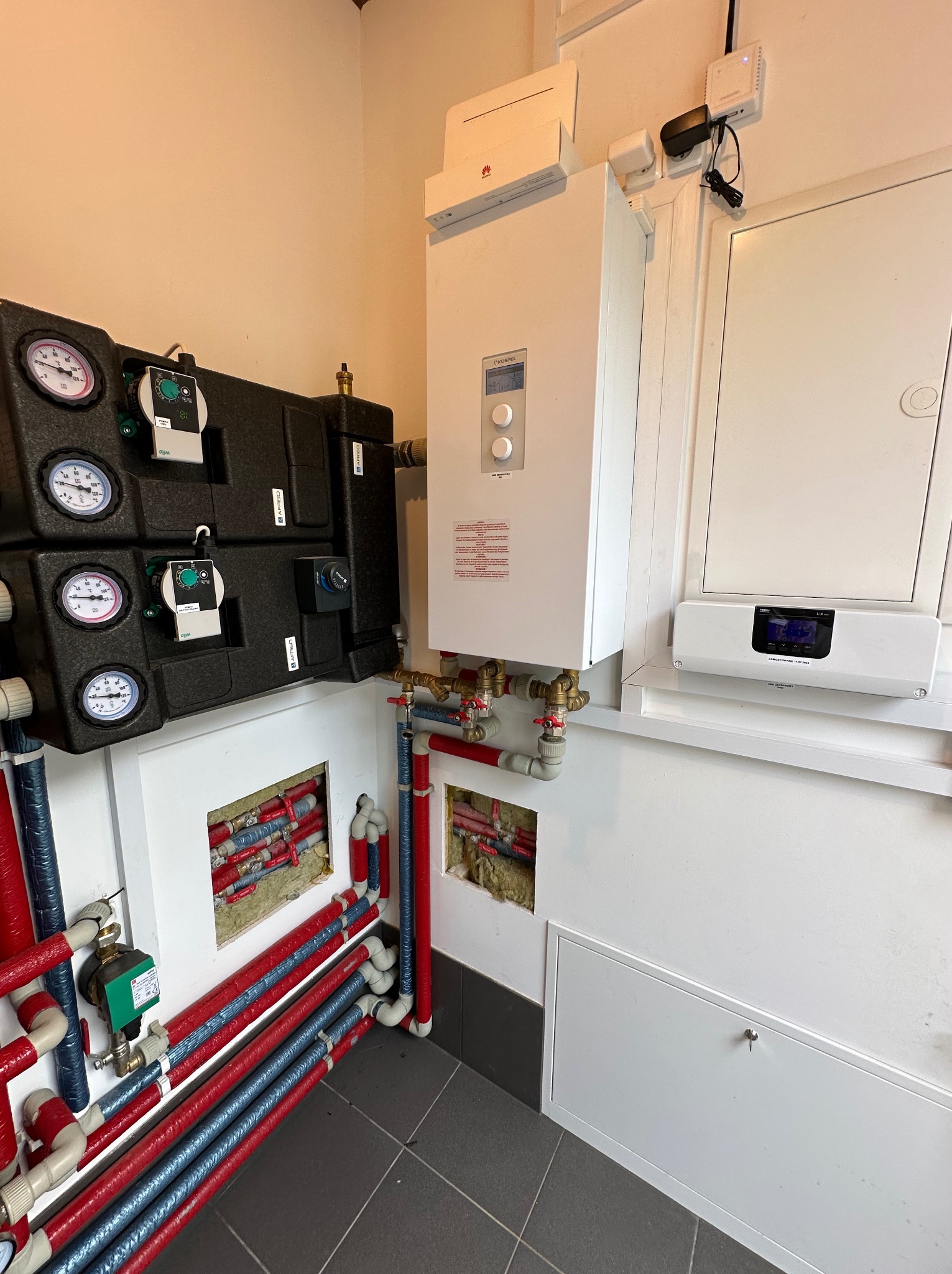 Węzeł cieplny z dwoma obiegami grzewczymi, widoczne pompy obiegowe, manometry, zawory oraz rury w izolacji czerwono-niebieskiej. Na ścianie biały piec grzewczy i regulator temperatury.