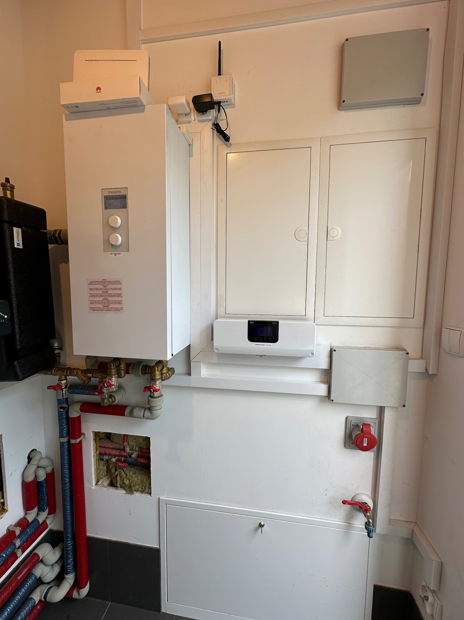 Biała instalacja grzewcza Kospel z widocznymi rurami, zaworami, czarnym zbiornikiem, skrzynkami elektrycznymi i routerem Huawei na górze, zamontowana na białej ścianie w pomieszczeniu technicznym.