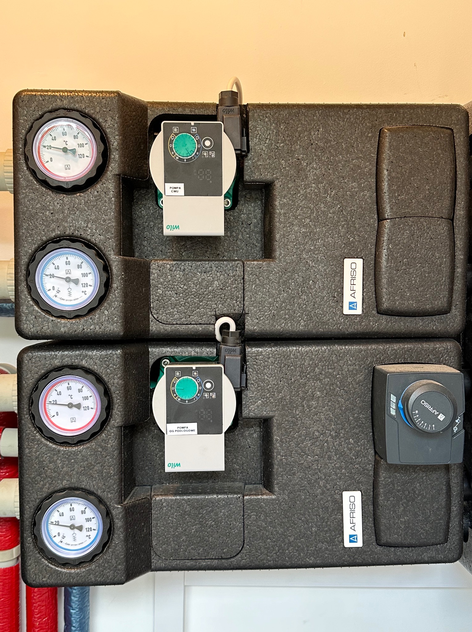 Dwie czarne, izolowane skrzynki z manometrami i regulatorami Wilo i Afriso, podłączone do czerwonych i niebieskich rur, prawdopodobnie element instalacji grzewczej lub klimatyzacji.
