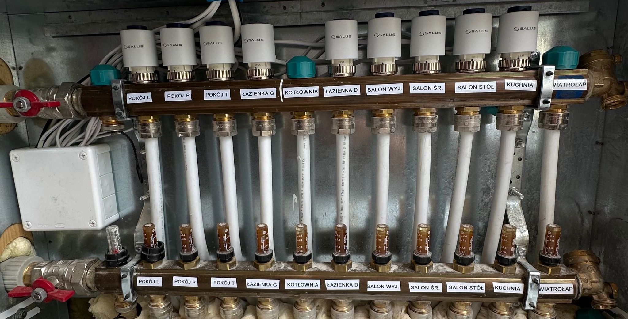 Rozdzielacz ogrzewania podłogowego z termostatami Salus, oznaczony dla różnych pomieszczeń: pokój L, P, T, łazienka G, kotłownia, łazienka D, salon WYJ., salon ŚR., salon STÓŁ, kuchnia, wiatrołap.