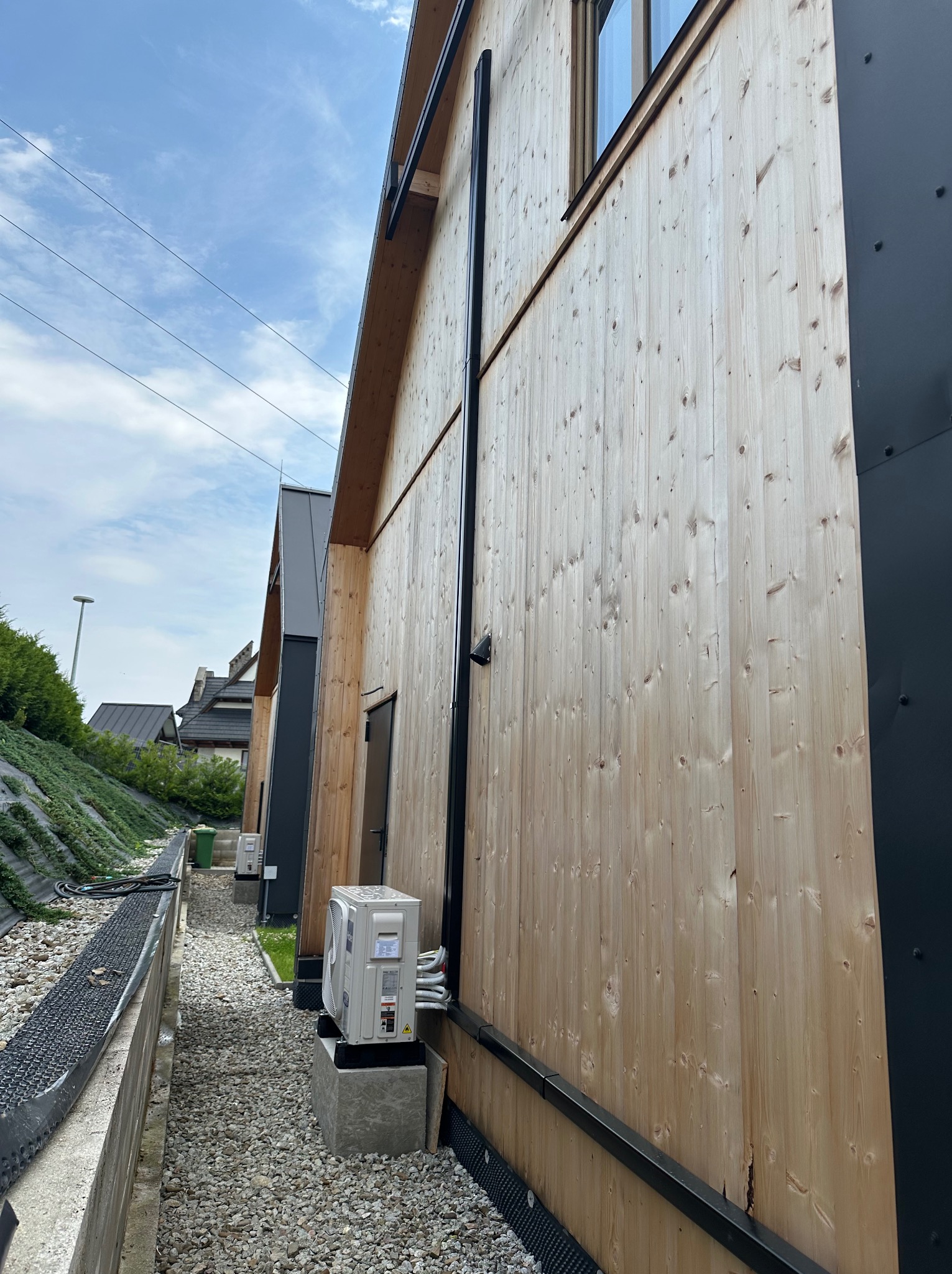 Zewnętrzna jednostka klimatyzacji typu split zamontowana na betonowym fundamencie przy drewnianej elewacji budynku, z widocznymi przewodami i rurami odprowadzającymi skropliny.