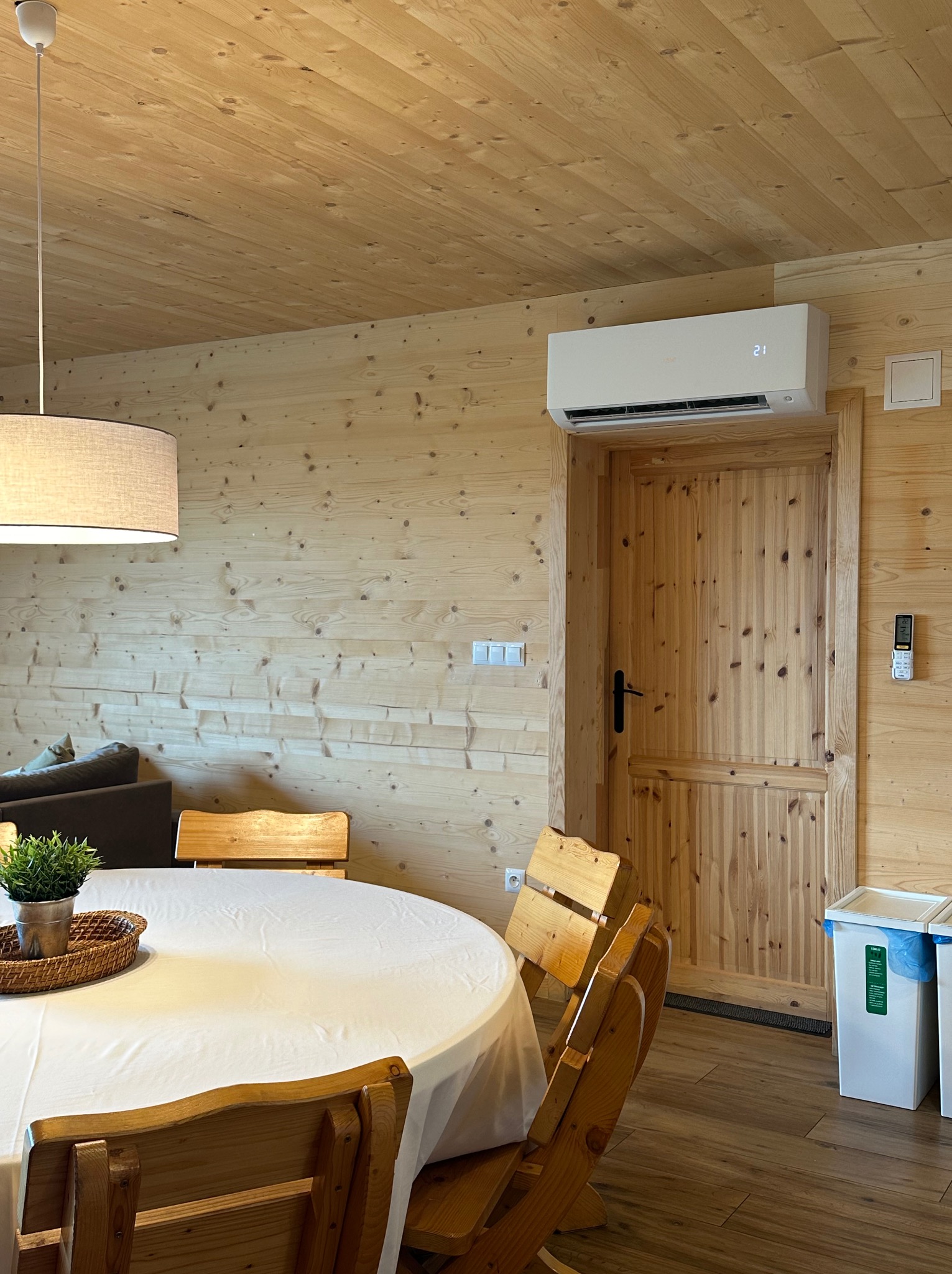 Wnętrze drewnianego domu z zamontowaną klimatyzacją nad drewnianymi drzwiami, widoczny okrągły stół z białym obrusem i drewniane krzesła.