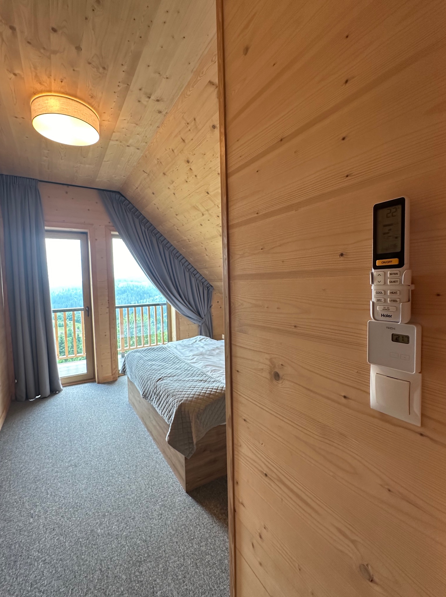 Drewniana sypialnia na poddaszu z widokiem na góry, klimatyzator Haier z pilotem na ścianie i szarymi zasłonami.