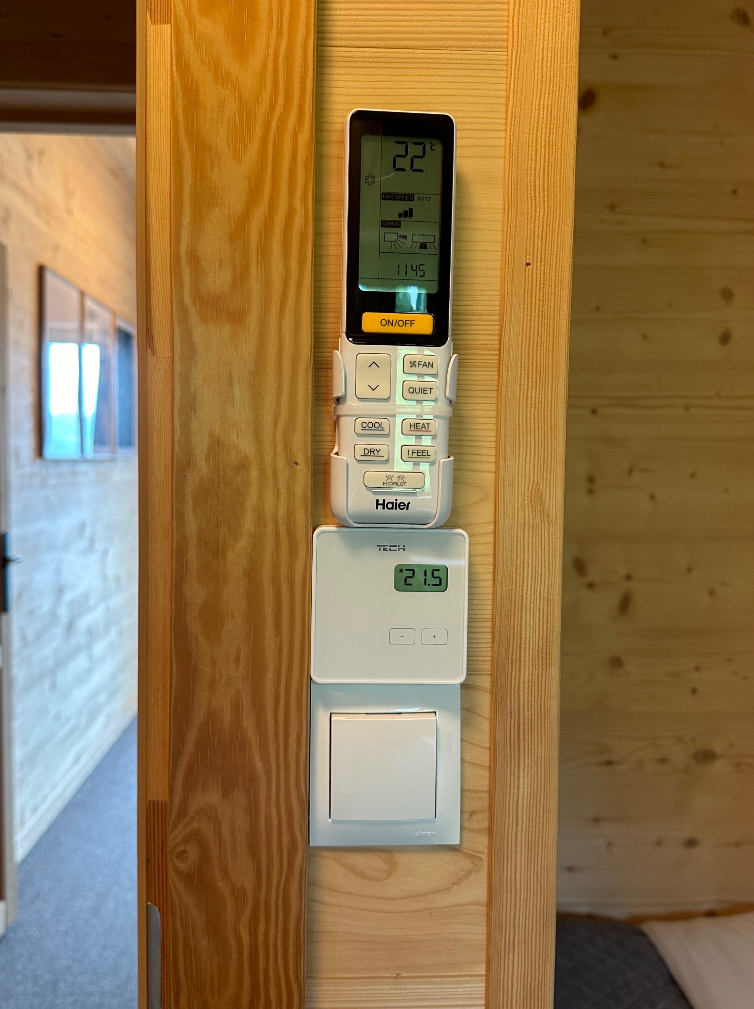 Panel sterowania klimatyzacją Haier z pilotem, termostat TECH i włącznik światła Simon zamontowane na drewnianej ścianie w domu, ustawiona temperatura 22 stopnie Celsjusza.