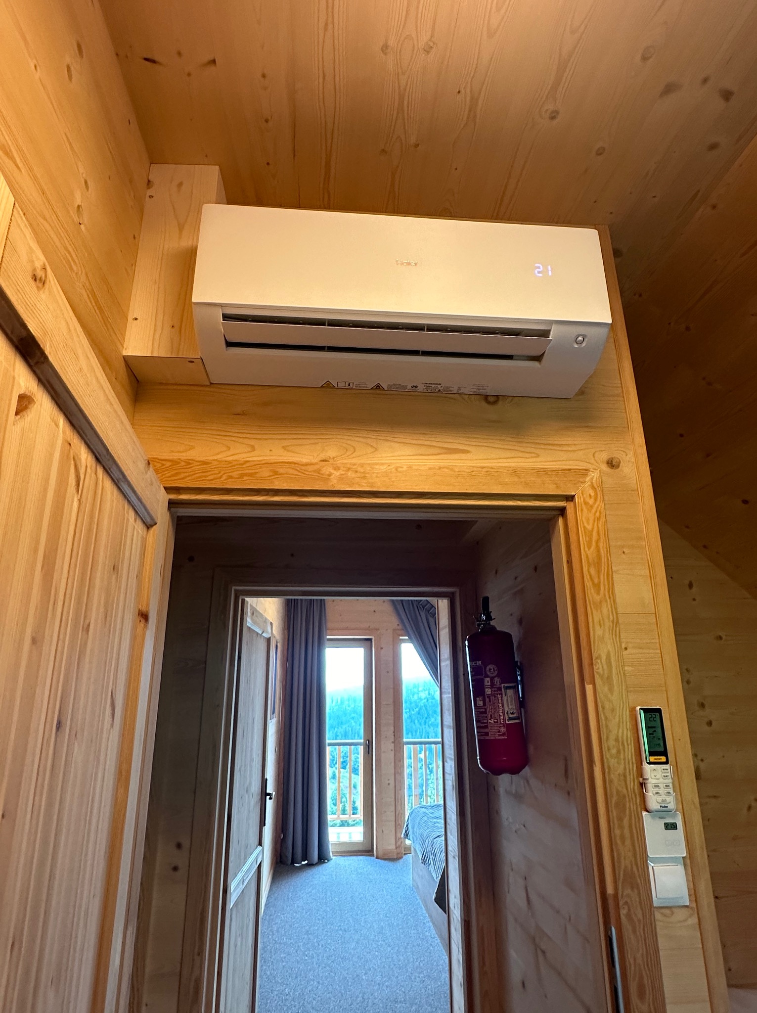 Biały klimatyzator Haier zamontowany na drewnianej ścianie w pokoju z boazerią, widok z dołu na urządzenie, z widocznym wyświetlaczem temperatury i fragmentem pokoju z balkonem w tle.