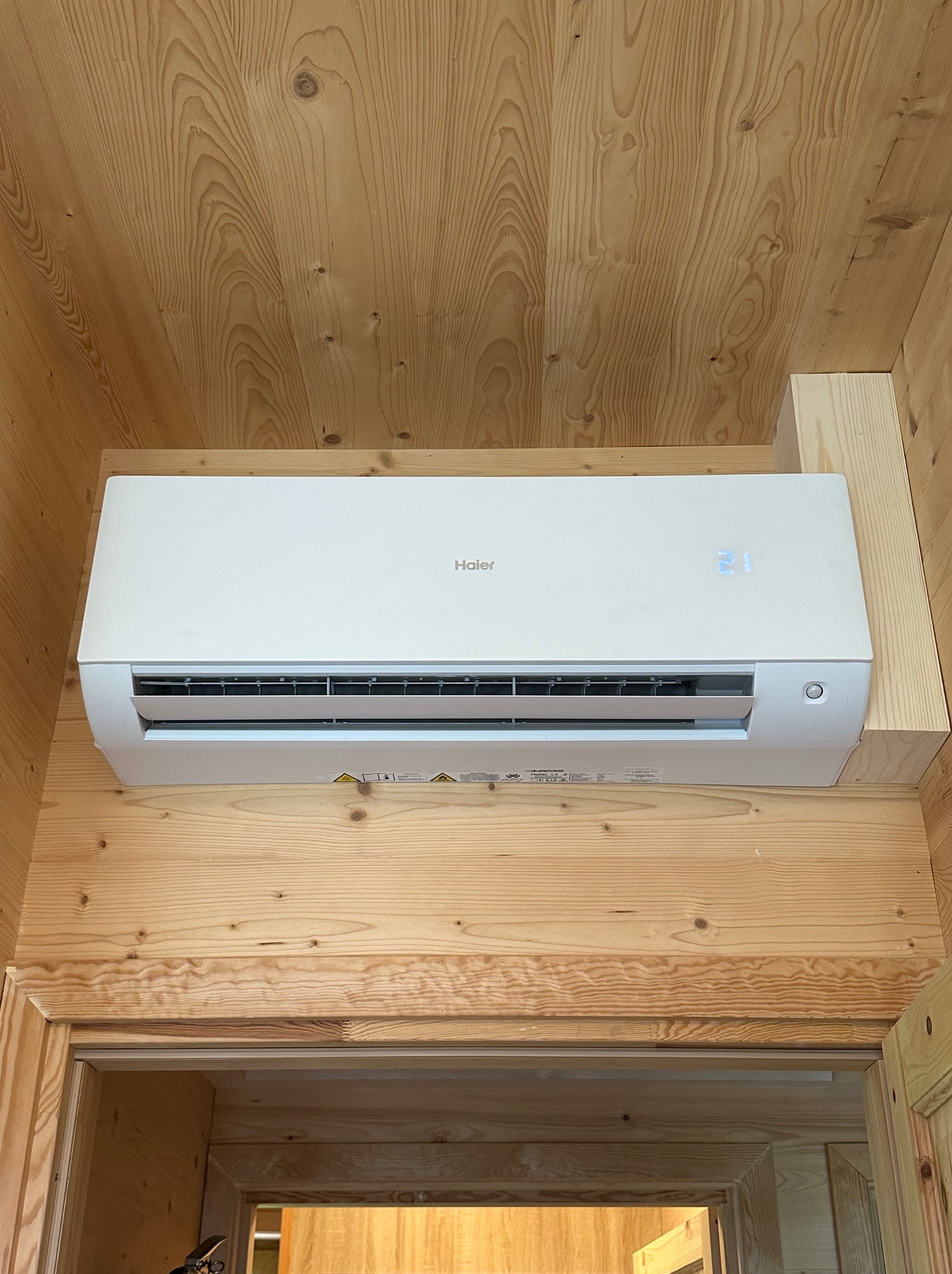 Biały klimatyzator Haier zamontowany na drewnianej ścianie wewnątrz pomieszczenia, widoczny wyświetlacz temperatury 21 stopni Celsjusza.