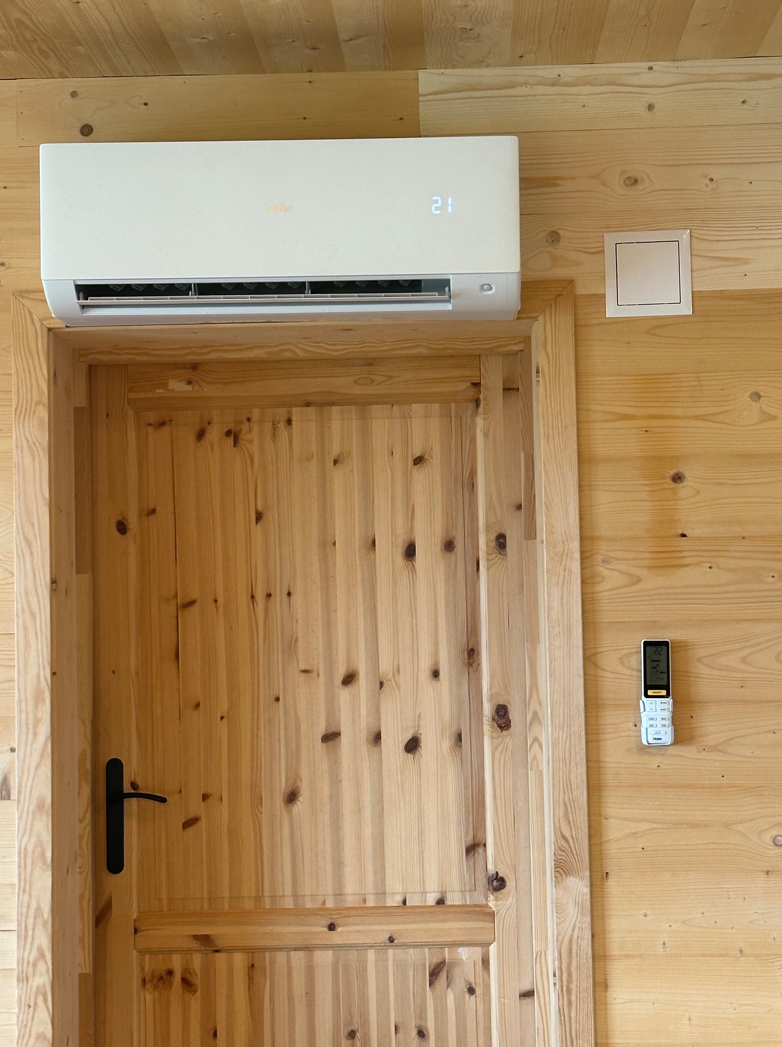 Biała klimatyzacja marki Haier zamontowana nad drewnianymi drzwiami w pomieszczeniu wykończonym boazerią, z widocznym panelem sterowania i pilotem na ścianie.