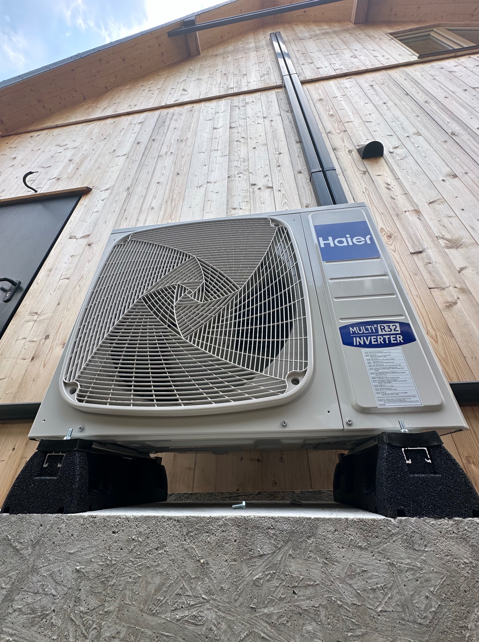 Zewnętrzna jednostka klimatyzacji Haier Multi R32 Inverter zamontowana na betonowym fundamencie przy drewnianej ścianie budynku, widok z dołu.