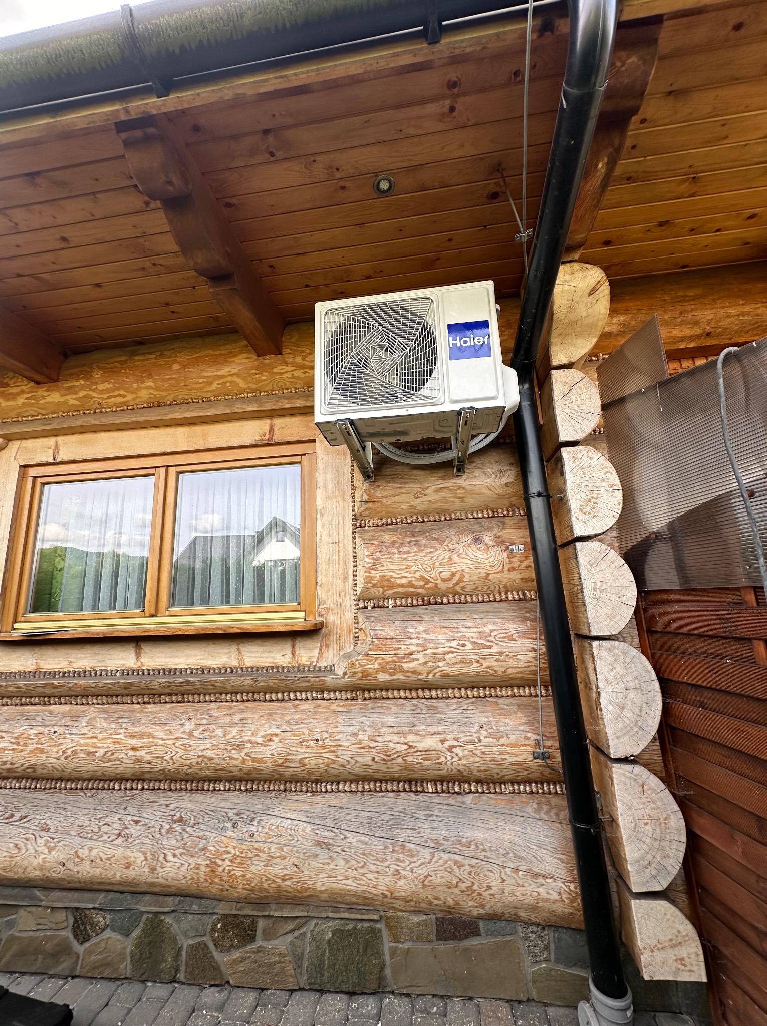 Biała jednostka zewnętrzna klimatyzacji Haier zamontowana na drewnianej ścianie domu z bali, widoczna rynna spustowa i okno z firankami.