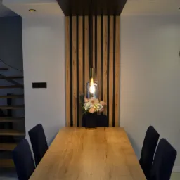 Jadalnia z drewnianym stołem, czterema granatowymi krzesłami i ozdobnym oświetleniem wiszącym nad stołem, na tle ściany z drewnianymi panelami.