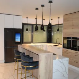 Nowoczesna kuchnia z wyspą, czarnymi lampami wiszącymi i sprzętem AGD w zabudowie, widok na marmurowe podłogi i welurowe hokery z złotymi nogami.