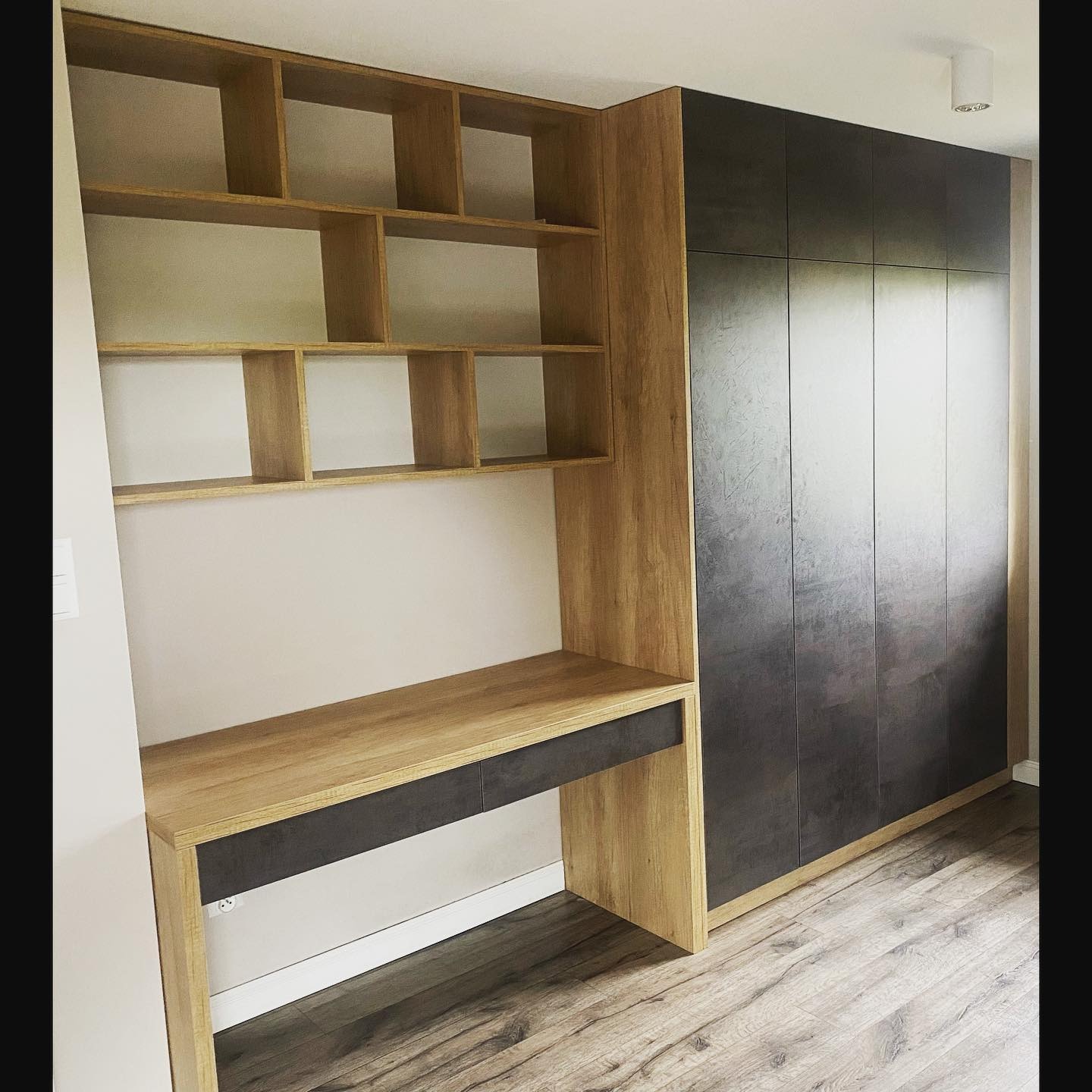 Zabudowa wnęki z biurkiem i szafą z ciemnoszarymi frontami, drewnopodobne wykończenie, widoczne półki nad biurkiem i drewniana podłoga.