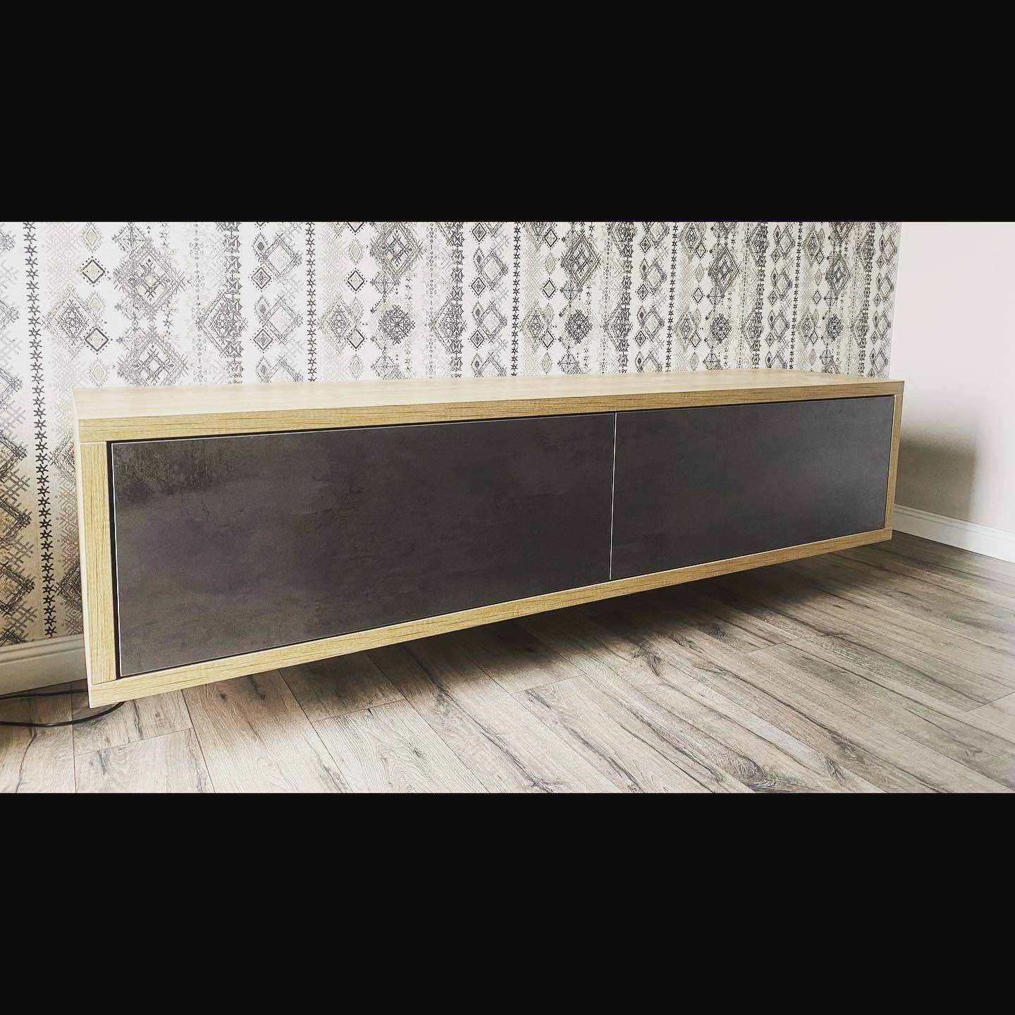 Nowoczesna szafka RTV wisząca, z jasną drewnianą ramą i ciemnymi, matowymi frontami, na tle wzorzystej tapety i drewnopodobnej podłogi.
