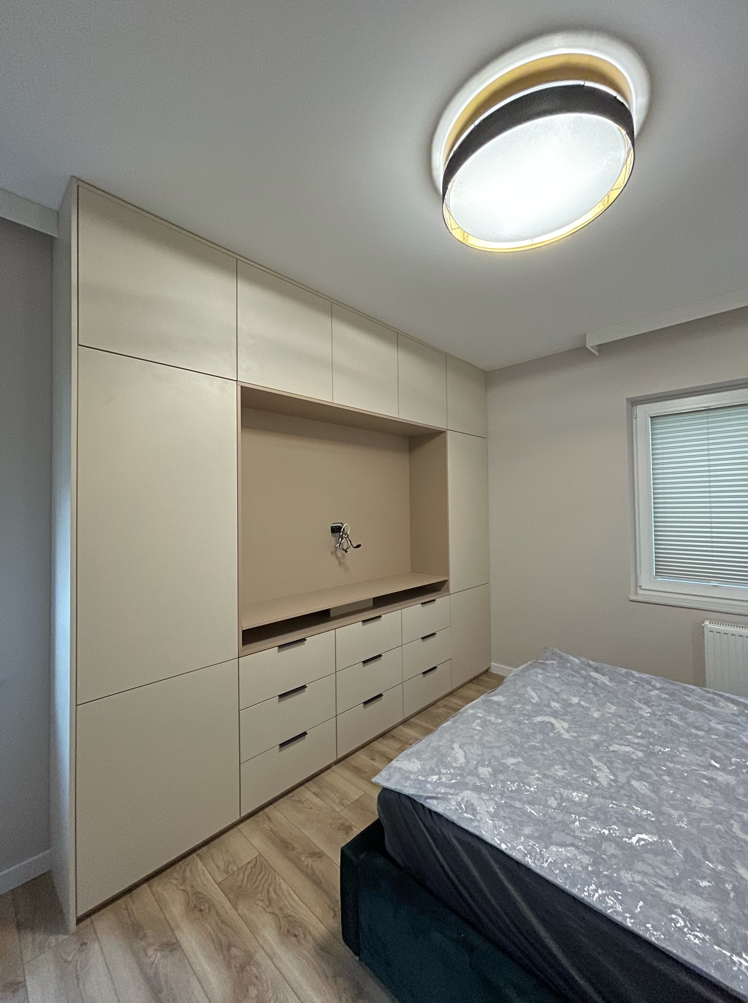 Beżowa zabudowa meblowa z szufladami i otwartą wnęką na ścianie w sypialni, widoczne gniazdko elektryczne, parkiet i fragment łóżka.