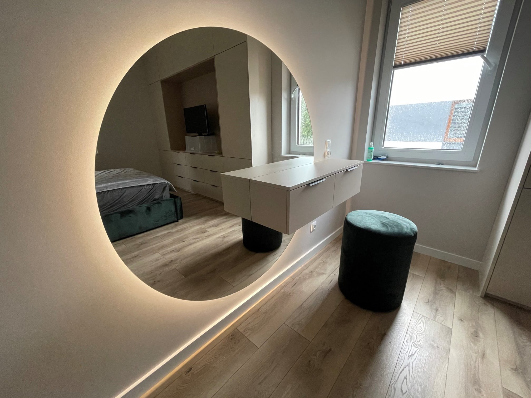 Nowoczesna sypialnia z toaletką z szufladami, okrągłym lustrem z podświetleniem LED i tapicerowanym pufem w kolorze butelkowej zieleni, odbicie łóżka i szafy w lustrze, jasna podłoga drewniana.
