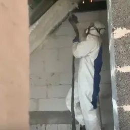 Pracownik w kombinezonie ochronnym i masce aplikuje piankę izolacyjną na poddaszu, stojąc na drabinie. Widoczna surowa ściana z bloczków i elementy konstrukcji dachu.