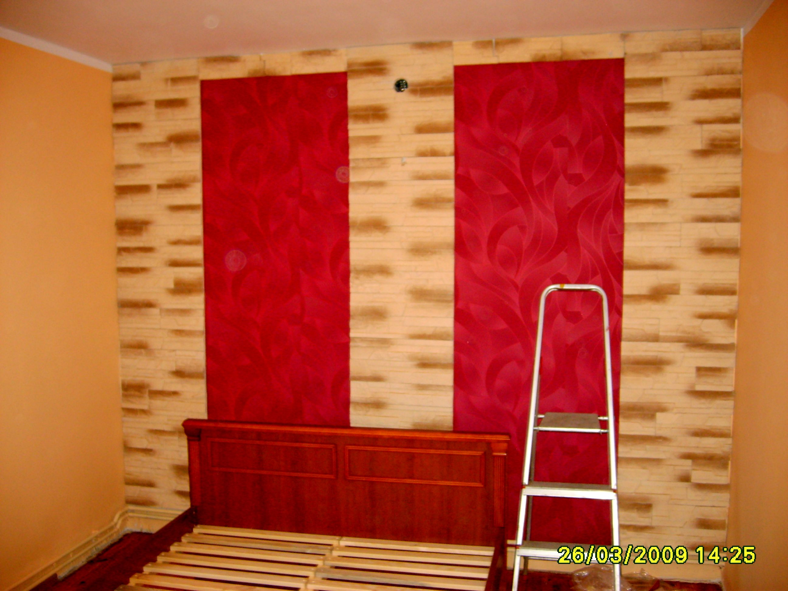 Wnętrze pokoju w trakcie remontu: ściany z dekoracyjnym tynkiem imitującym cegłę i czerwonymi panelami z wzorem, widoczna drabina i rama łóżka bez materaca.