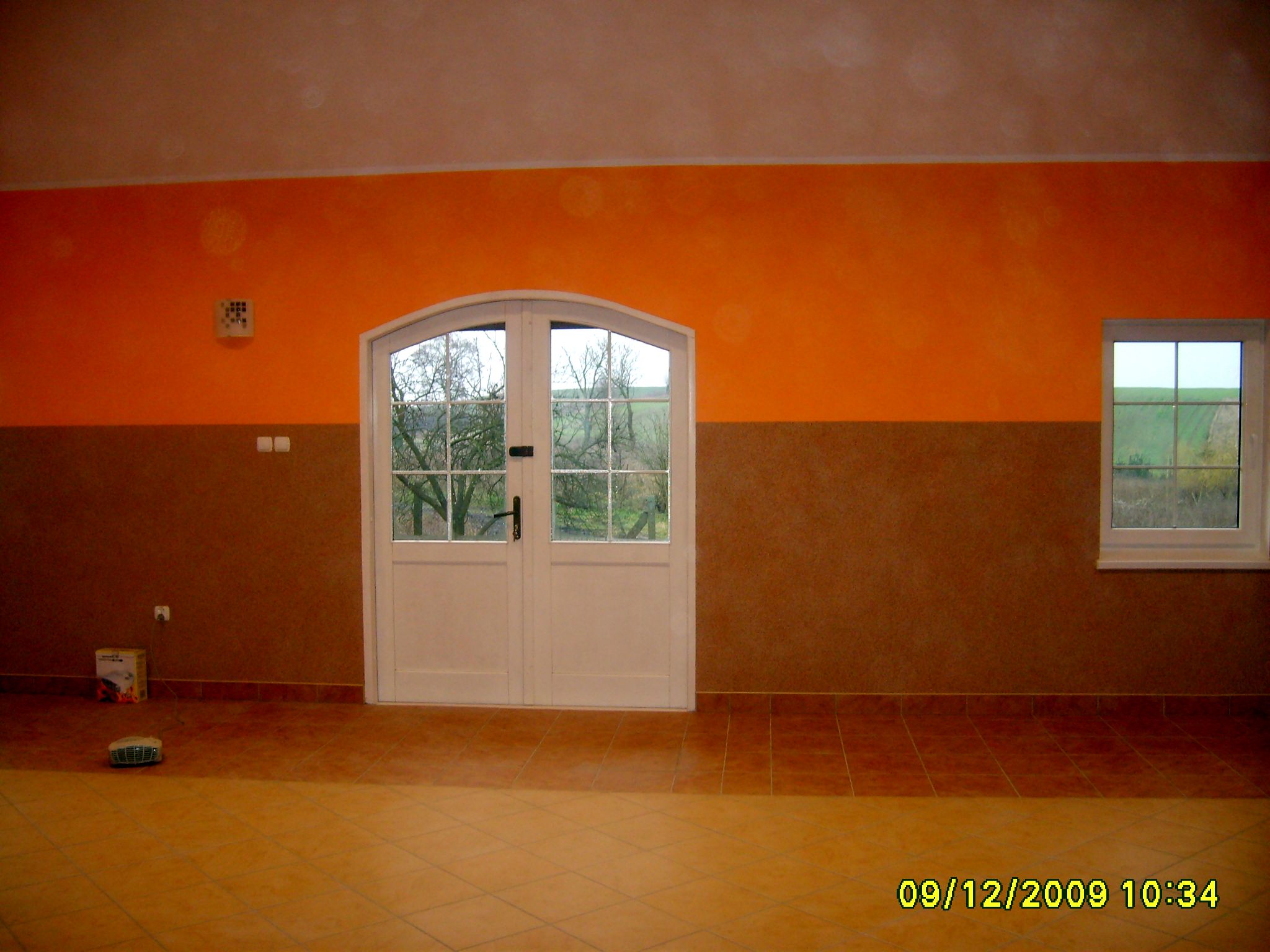 Wnętrze po remoncie: ściana pomalowana na pomarańczowo i brązowo, białe dwuskrzydłowe drzwi z szybami wychodzące na zewnątrz, okno z widokiem na zieleń, brązowa podłoga z płytek.