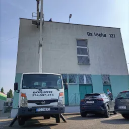 Pracownik na podnośniku koszowym przy budynku Os. Lecha 121, firma BORKOSZ z numerem 574-597-914, zaparkowane samochody osobowe w tle.