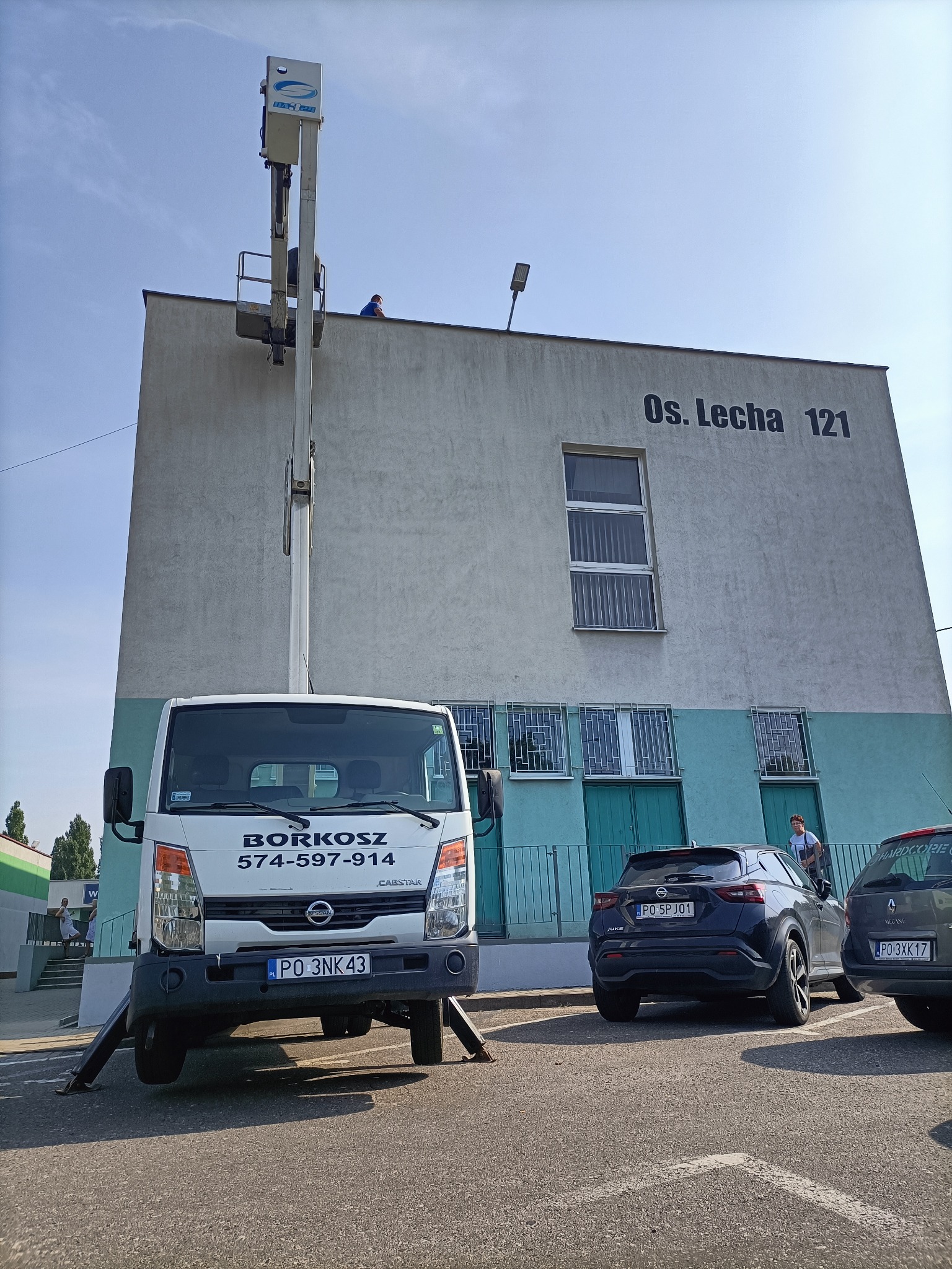 Pracownik na podnośniku koszowym przy budynku Os. Lecha 121, firma BORKOSZ z numerem 574-597-914, zaparkowane samochody osobowe w tle.