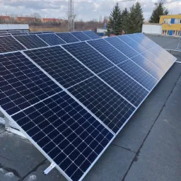 Rząd paneli słonecznych na płaskim dachu, z widocznymi mocowaniami i elementami konstrukcyjnymi, w tle budynki i drzewa pod pochmurnym niebem.