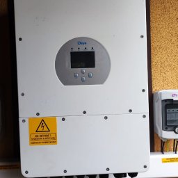 MR.OZE KONRAD ROZDOBA - Biały inwerter firmy Deye zamontowany na ścianie o pomarańczowej strukturze, z widocznymi kablami i żółtą naklejką ostrzegawczą.