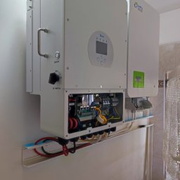 MR.OZE KONRAD ROZDOBA - Dwa białe inwertery zamontowane na ścianie w pomieszczeniu, widoczne przewody, otwarte skrzynki z elektroniką, z lewej inwerter marki Deye z wyświetlaczem i wyłącznikiem DC, z prawej inwerter marki...