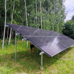 MR.OZE KONRAD ROZDOBA - Instalacja paneli słonecznych na konstrukcji wsporczej, widoczne stopy mężczyzny stojącego pod panelami, w tle las brzozowy.