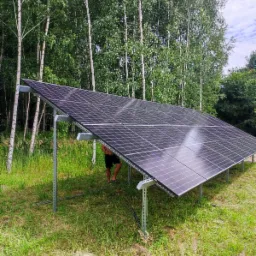 Instalacja paneli słonecznych na konstrukcji wsporczej, widoczne stopy mężczyzny stojącego pod panelami, w tle las brzozowy.