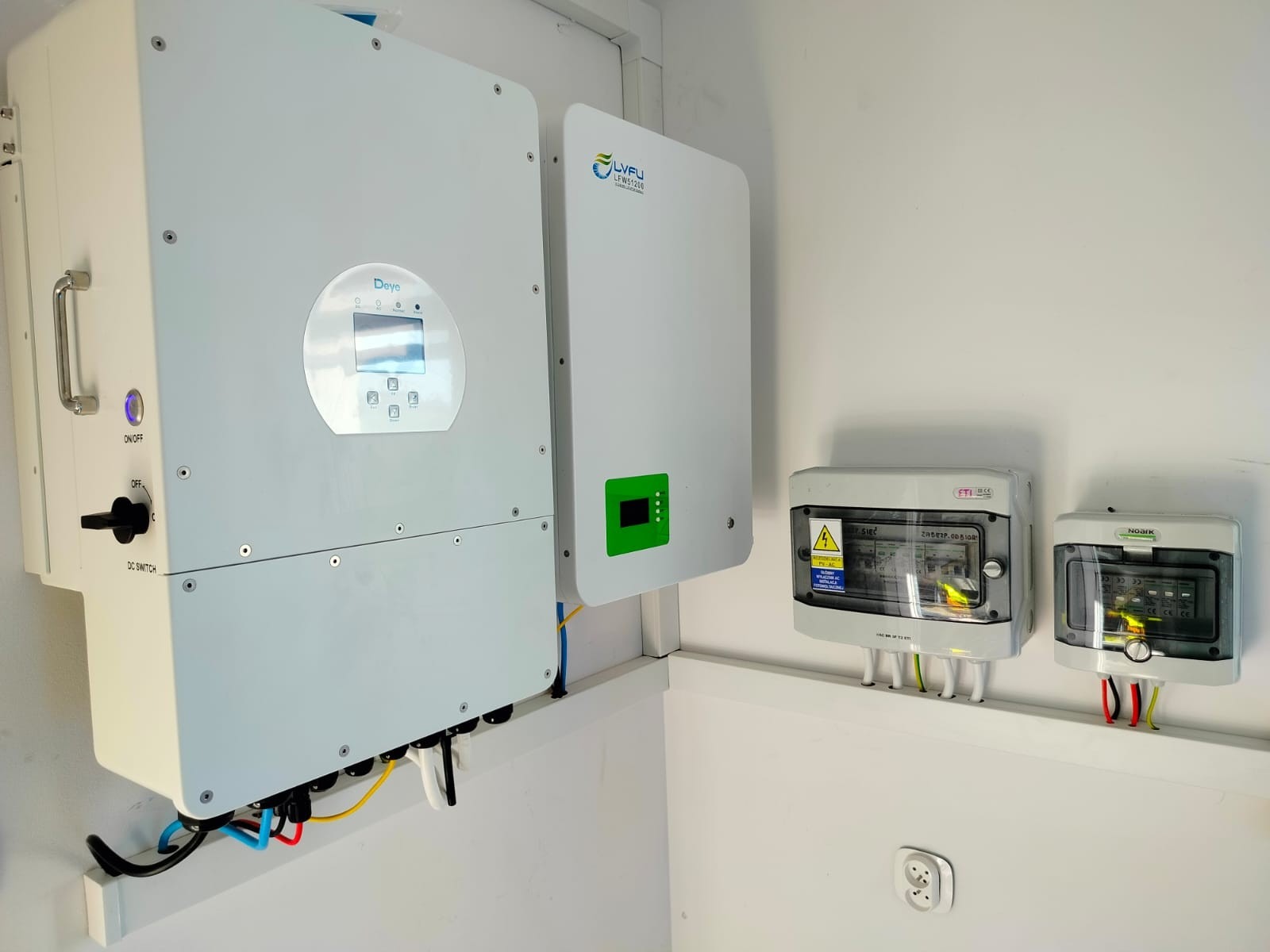 Dwa białe inwertery różnych marek (Deye i LYFU) zamontowane na białej ścianie obok dwóch skrzynek bezpiecznikowych z widocznymi kablami.