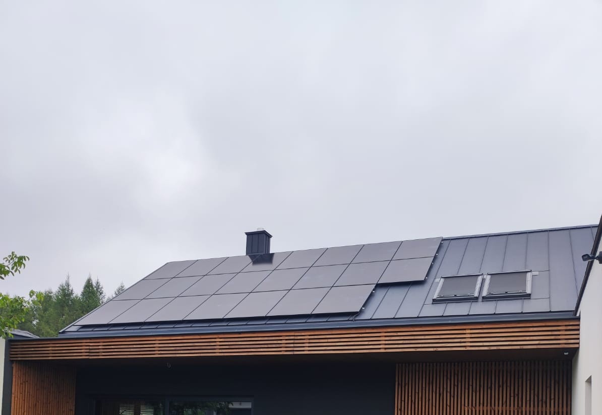 Ciemne panele słoneczne na dachu domu z blachy, widoczne dwa okna dachowe i komin, pochmurne niebo w tle.