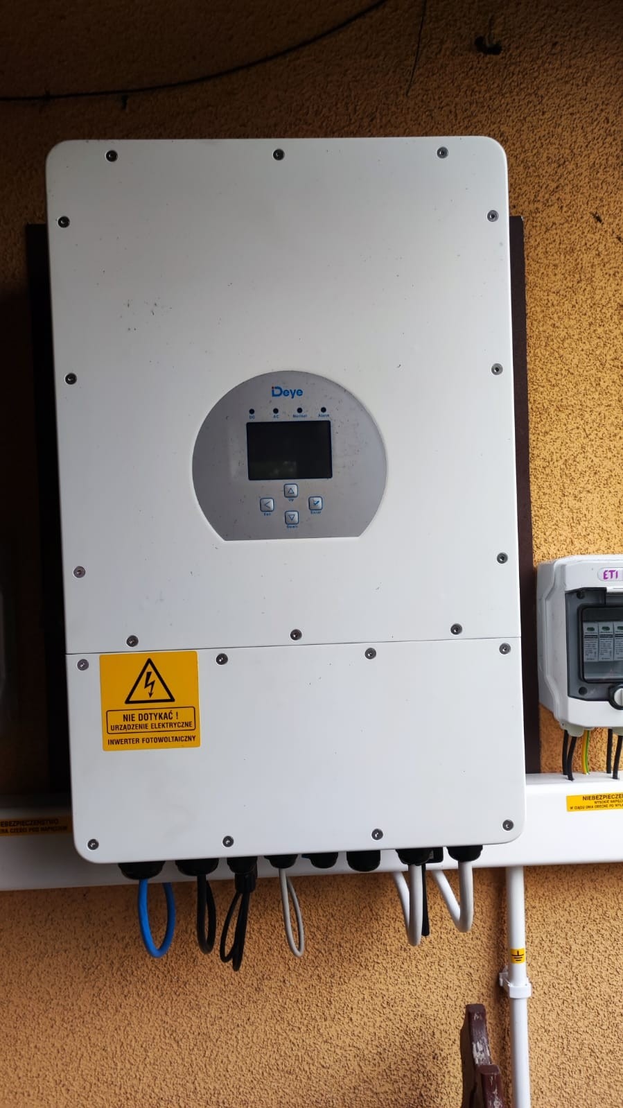 Biały inwerter firmy Deye zamontowany na ścianie o pomarańczowej strukturze, z widocznymi kablami i żółtą naklejką ostrzegawczą.