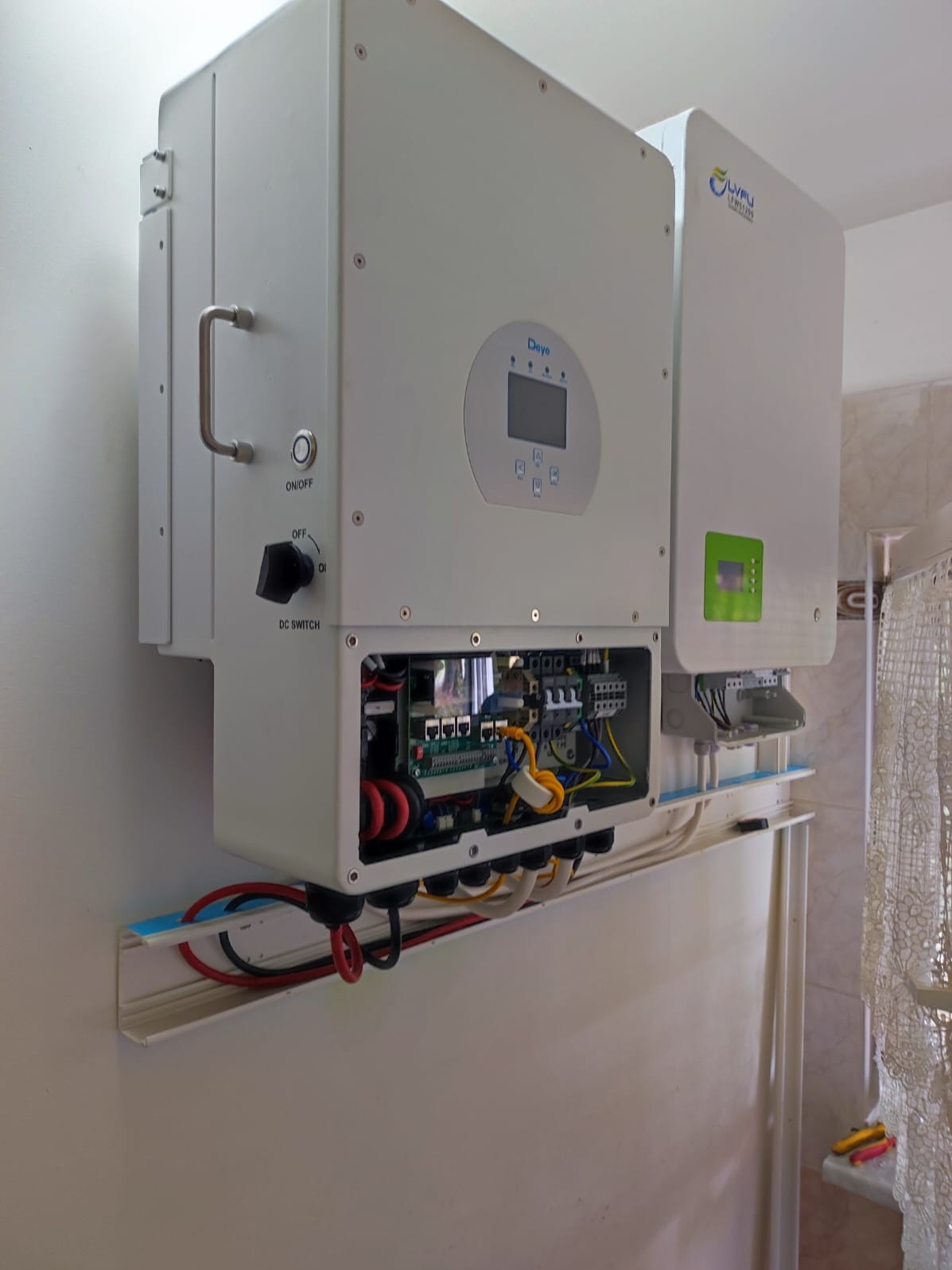 Dwa białe inwertery zamontowane na ścianie w pomieszczeniu, widoczne przewody, otwarte skrzynki z elektroniką, z lewej inwerter marki Deye z wyświetlaczem i wyłącznikiem DC, z prawej inwerter marki...