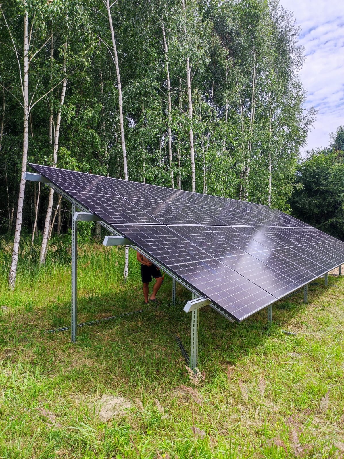 Instalacja paneli słonecznych na konstrukcji wsporczej, widoczne stopy mężczyzny stojącego pod panelami, w tle las brzozowy.