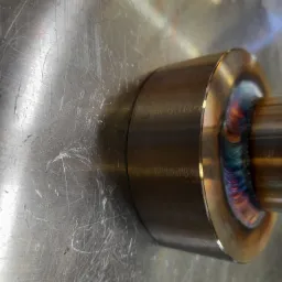 Detale spoiny na metalowej rurze z widocznymi kolorami hartowania, leżącej na porysowanej, metalowej powierzchni.